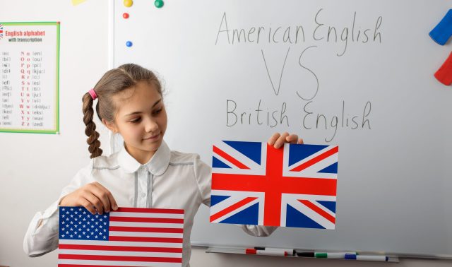 American English VS British English concept. Cheerful schoolgirl with American and British flags in the classroom, learning differences in types of English languages